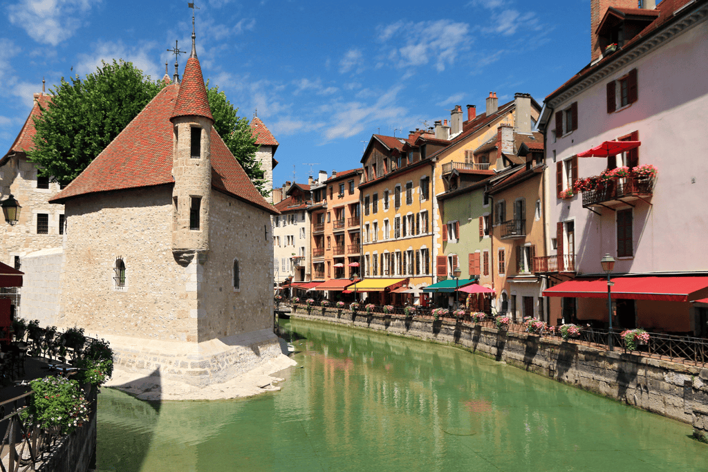 Palais de l Ile sits in the Thiou canal in Annecy surrounded by colorful historic buildings with flower boxes and cafe awnings. The picturesque old town is a popular day trip from Lyon and a scenic addition to a Lyon itinerary.