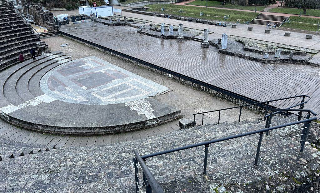 Stone seating overlooks the ancient Roman theater in Lyon with a semicircular mosaic floor and a wooden stage lined with weathered columns. The Roman ruins at Lugdunum are part of the historic fabric of Lyon and help illustrate why the city is recognized as a UNESCO World Heritage Site.