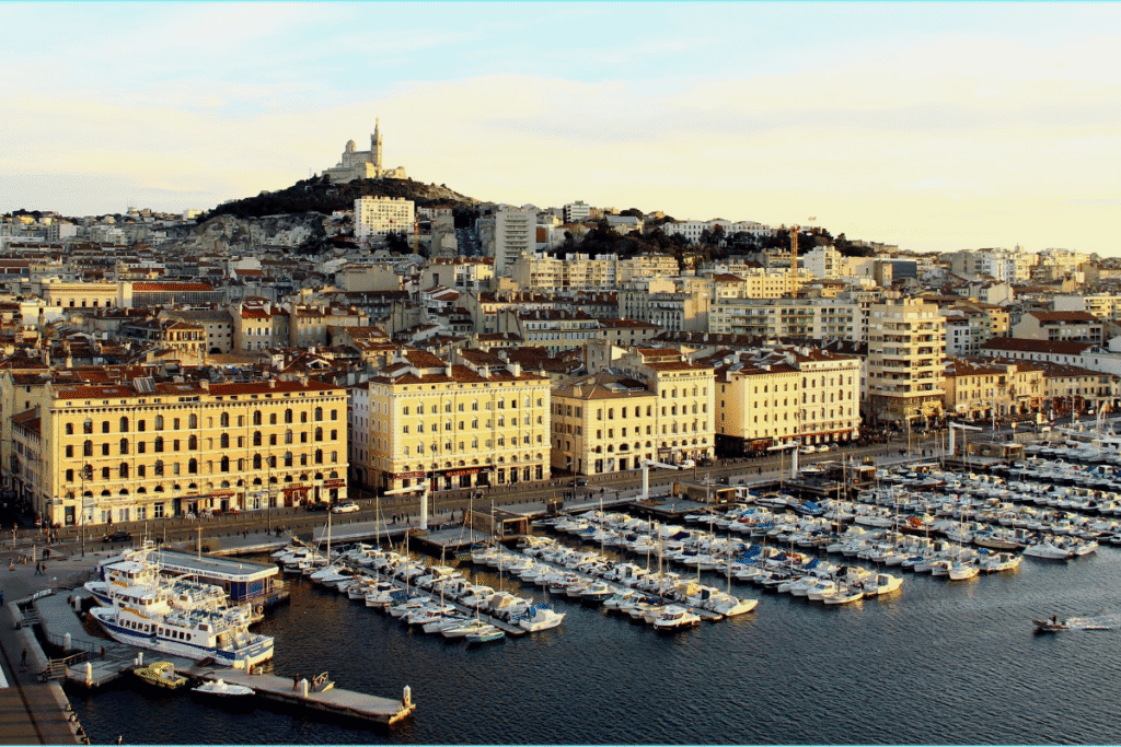 The Vieux Port of Marseille with boats and yachts docked along the harbor is shown in the foreground. Behind the historic waterfront buildings, the basilica of Notre Dame de la Garde stands on a hill overlooking the city. This iconic view highlights why Marseille is worth visiting.