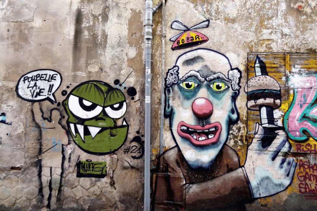 Colorful street art on a weathered wall in the Panier Distric in Marseille, featuring a green cartoonish face with sharp teeth and a speech bubble saying "Poubelle la vie (Trash Life in English)," alongside a clown-like figure holding a burger with a knife through it. This edgy, expressive graffiti scene reflects the urban creativity and alternative culture that make street art one of the intriguing things to see in Marseille.