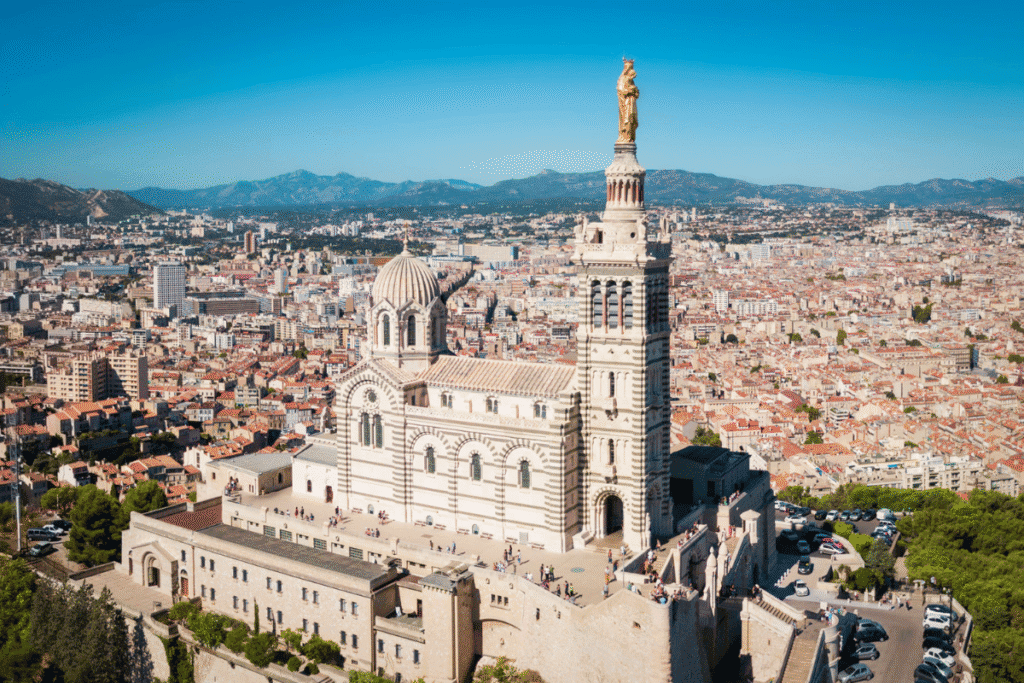 erial view of the Notre-Dame de la Garde basilica perched on a hilltop overlooking the city of Marseille, with its striped stone facade, domed roof, and golden statue of the Virgin Mary glistening under a clear blue sky. This iconic landmark is a must-see for anyone planning to travel to Marseille.