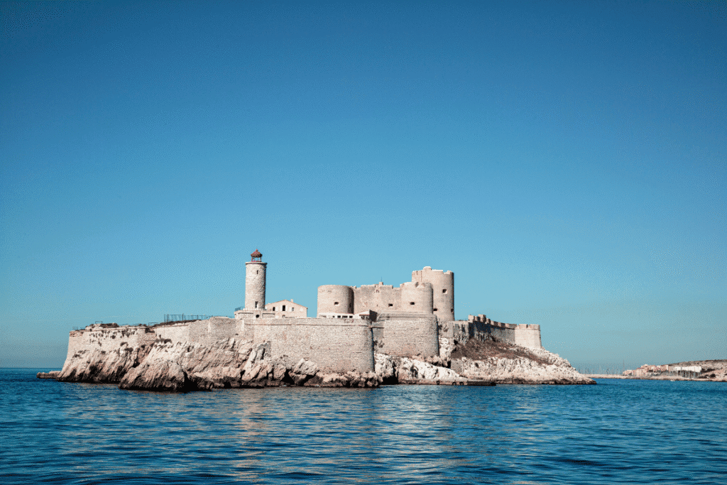Château d'If, a historic fortress on a rocky island off the coast of Marseille, stands against a clear blue sky and calm sea. The stone structure with its round towers and red-capped lighthouse evokes the city’s maritime history and literary fame, making it a compelling reason why Marseille is worth visiting.