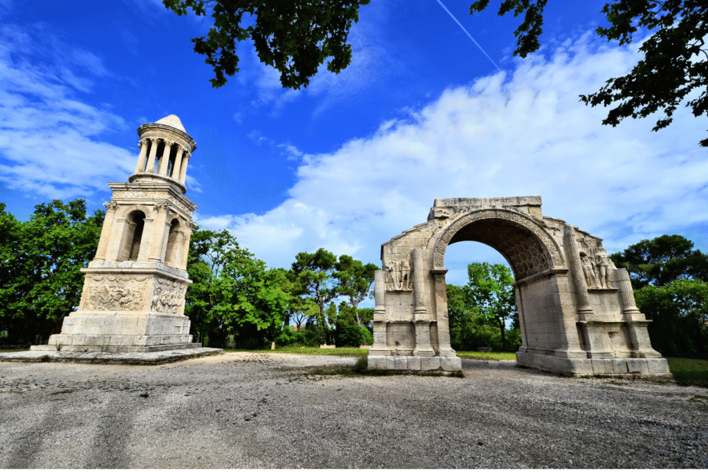 The Mausoleum of the Julii and the Triumphal Arch of Glanum, two remarkably preserved Roman ruins just outside Saint-Rémy-de-Provence, stand tall amid lush greenery under a bright blue sky. These ancient monuments are key highlights of a St Remy day trip from Marseille for history and archaeology enthusiasts.