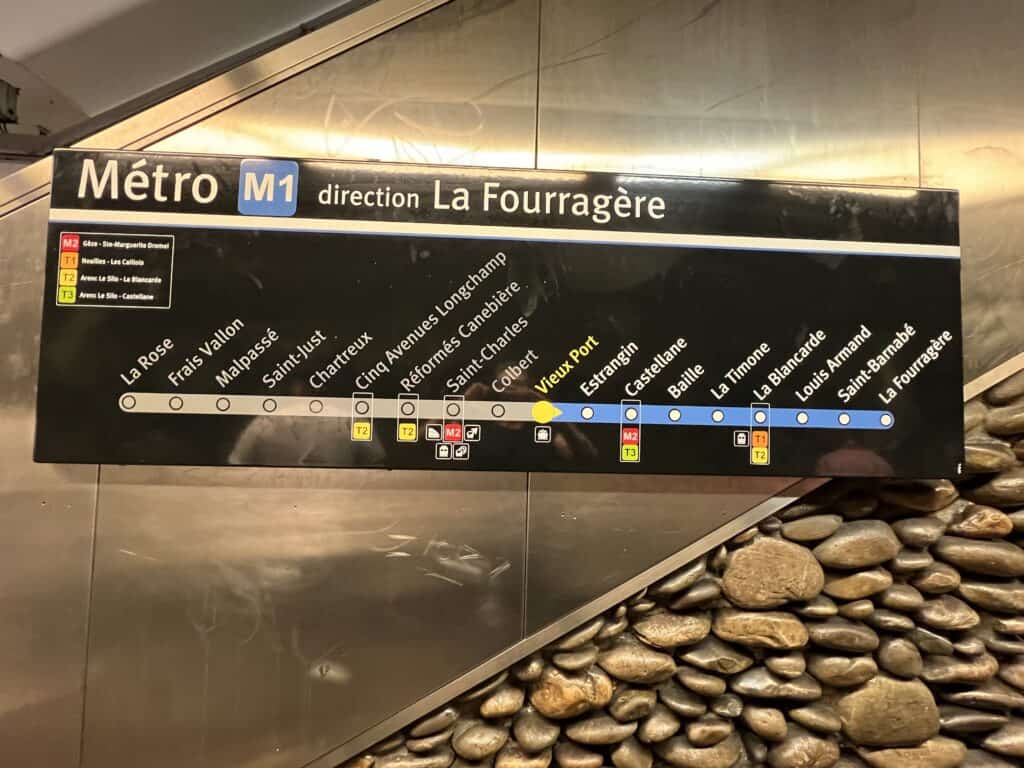 A sign for Marseille Metro Line M1 shows the route from La Rose to La Fourragère, with stops including Saint-Charles, Vieux Port, and Castellane. The highlighted Vieux Port station marks one of the most central spots for Marseille travel, connecting visitors to the city’s historic harbor and main attractions. The map is mounted on a wall with stone detailing beneath it.