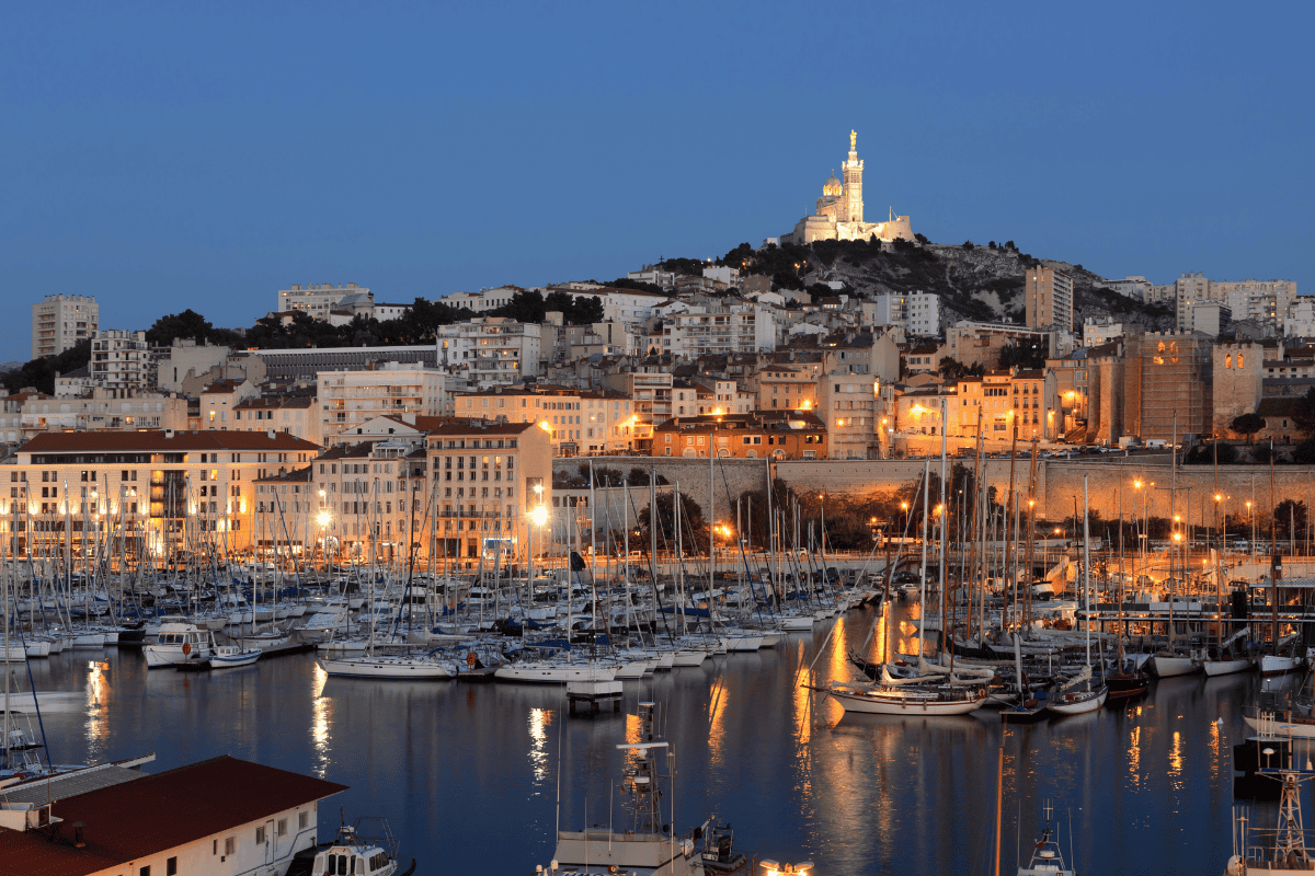 Evening view of Marseille’s Old Port with sailboats docked in the marina, city lights reflecting on the water, and the illuminated Basilica of Notre Dame de la Garde on the hilltop, a must-see stop on any Marseille itinerary.