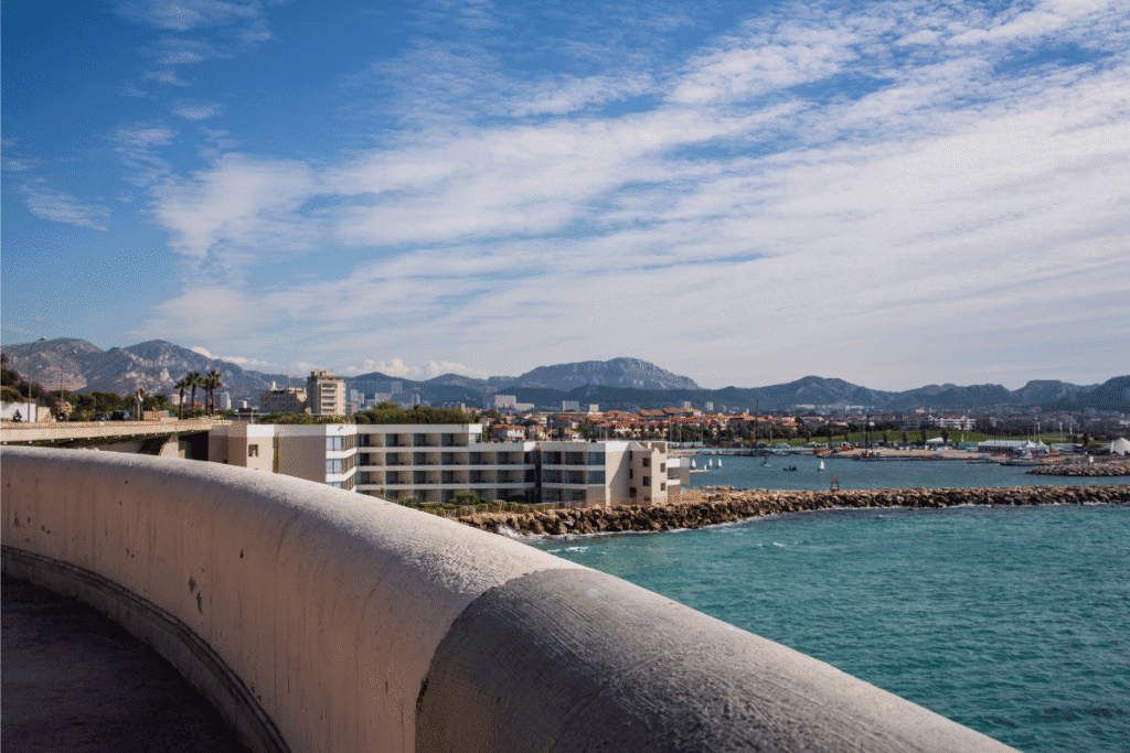 Scenic view from the Corniche Kennedy in Marseille, overlooking turquoise waters, modern buildings, and distant mountains under a partly cloudy sky, a picturesque stop along any Marseille itinerary.