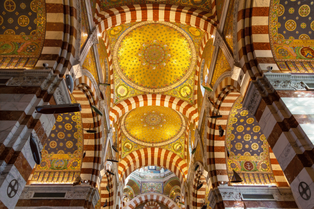 Ornate golden ceiling and striped arches inside the Basilica of Notre Dame de la Garde in Marseille, decorated with detailed mosaics and hanging model ships, a stunning highlight on any Marseille itinerary.