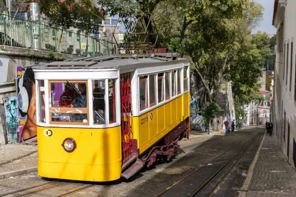 A classic yellow tram climbing a steep cobbled street lined with graffiti and greenery, capturing a quintessential moment from a Lisbon Itinerary.