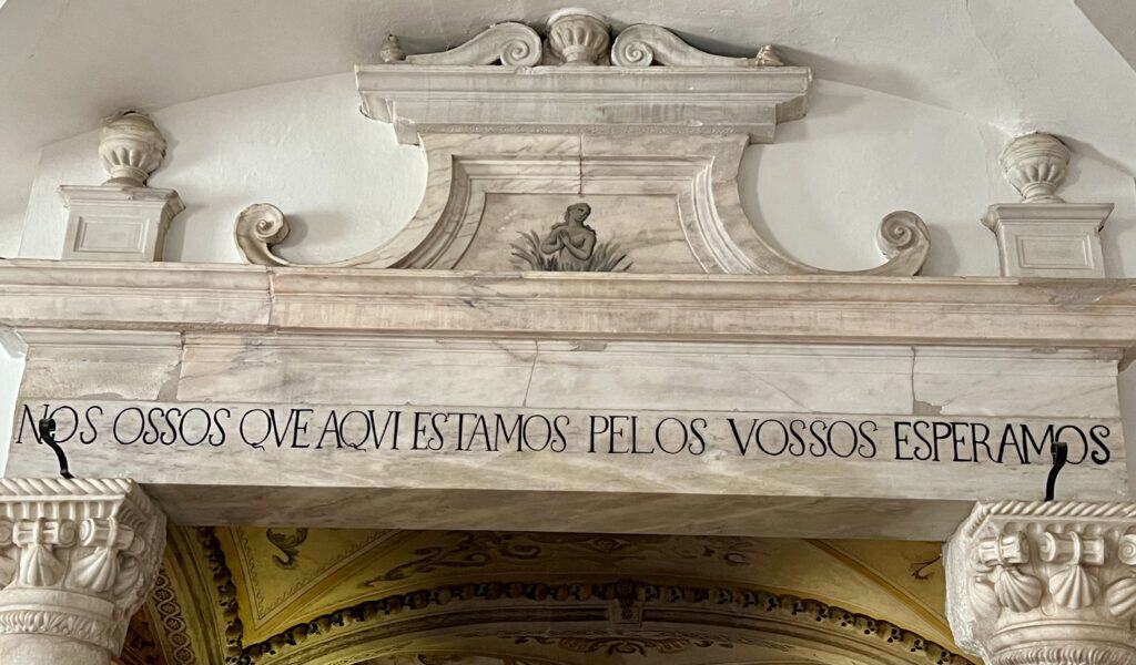 Stone archway inside the Chapel of Bones displays the inscription "NOS OSSOS QVE AQVI ESTAMOS PELOS VOSSOS ESPERAMOS" (which means " We bones that are here await yours" carved across the top. Decorative carvings and a small sculpted figure above the text highlight the eerie history often included in day trip to Evora from Lisbon.