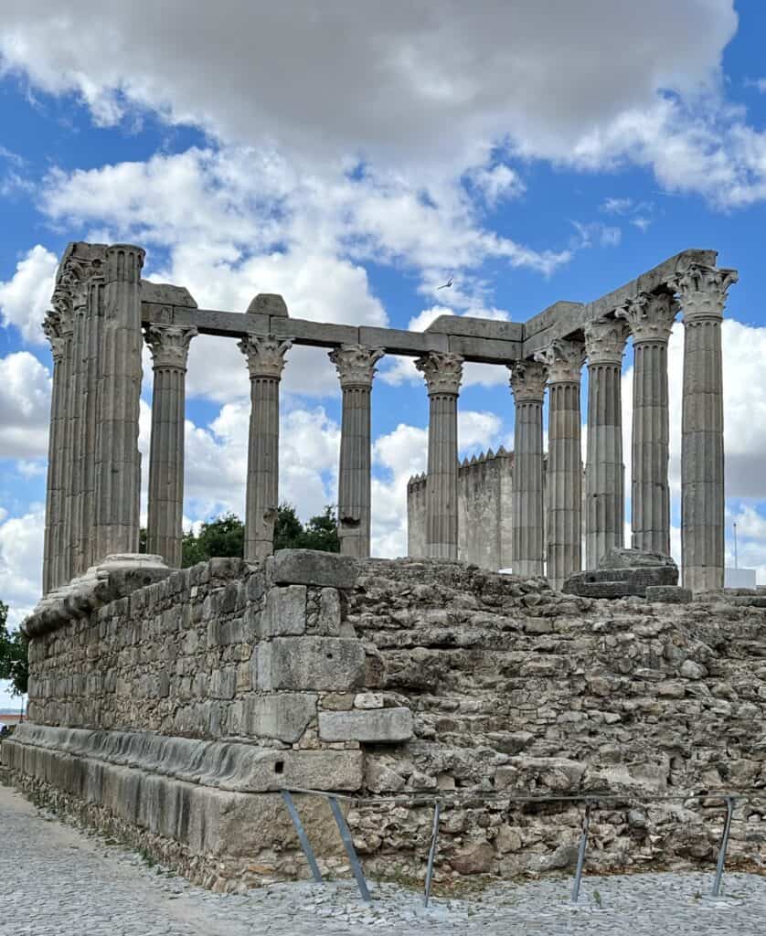 Ancient Roman temple ruins in Evora stand with tall stone columns against a bright sky with scattered clouds. This historic site is a popular option for day trips from Lisbon and adds depth to a longer Lisbon itinerary.