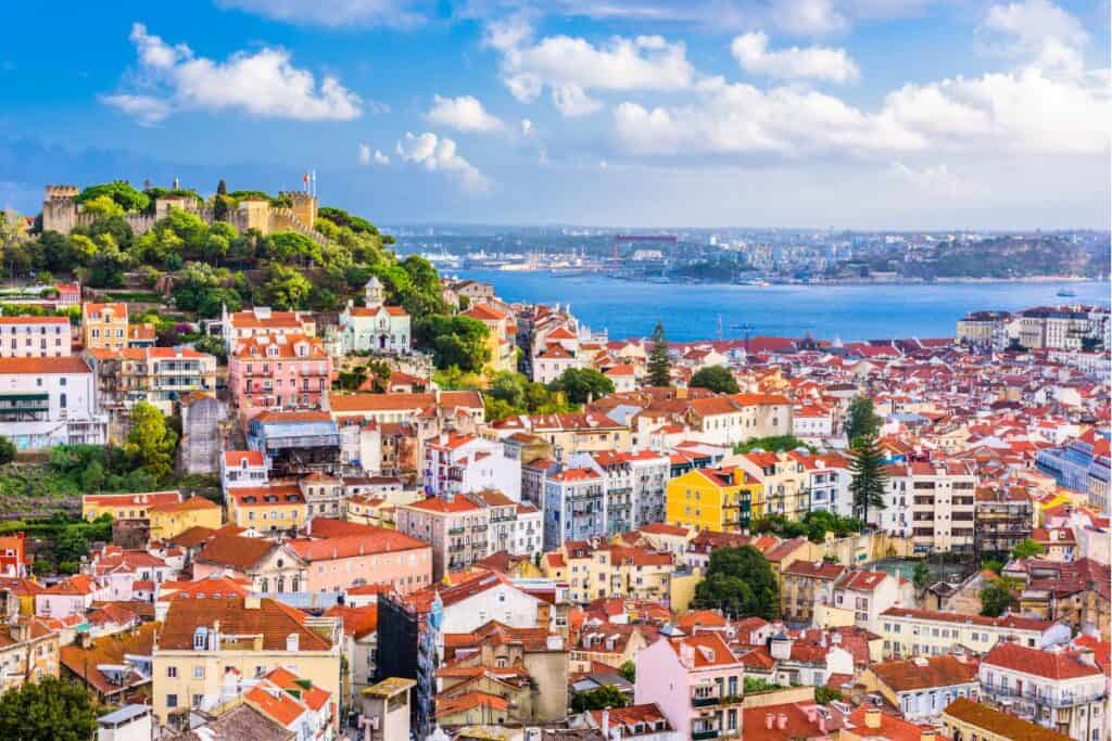 Panoramic view of Lisbon shows colorful buildings with red rooftops climbing a hillside toward a castle overlooking the Tagus River. This scenic viewpoint captures the charm of the city on a 3 day itinerary Lisbon.