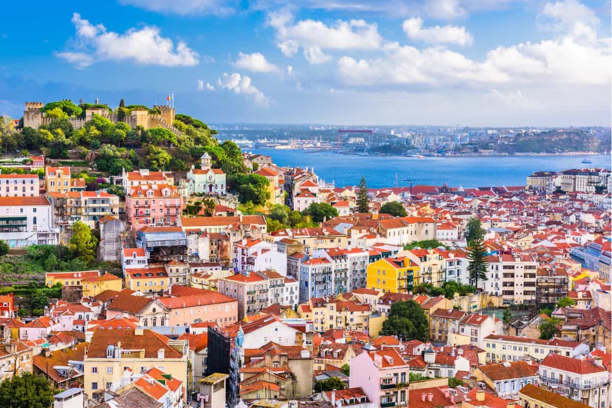 Panoramic view of Lisbon shows colorful buildings with red rooftops climbing a hillside toward a castle overlooking the Tagus River. This scenic viewpoint captures the charm of the city on a 3 day itinerary Lisbon.