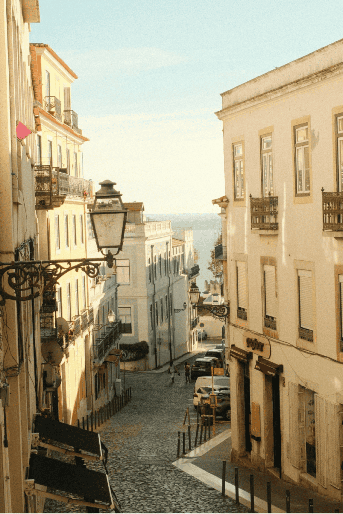 Narrow cobblestone street in the Alfama distric of Lisbon slopes downhill between pastel buildings with wrought iron balconies and vintage street lamps, opening toward the Tagus River in the distance. This scenic neighborhood view captures the charm of wandering through the city on a 3 day itinerary Lisbon.