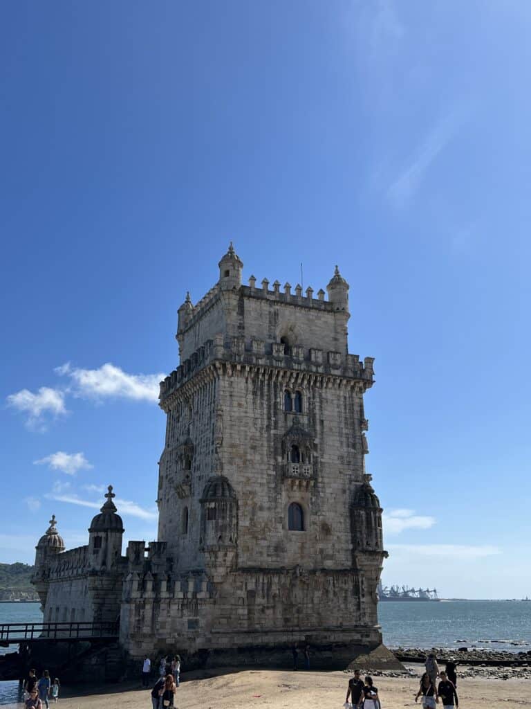 Belem Tower stands along the Tagus River with detailed stonework and small turrets under a clear blue sky while visitors walk nearby. This UNESCO World Heritage Site is a must see stop on a 3 day itinerary Lisbon.
