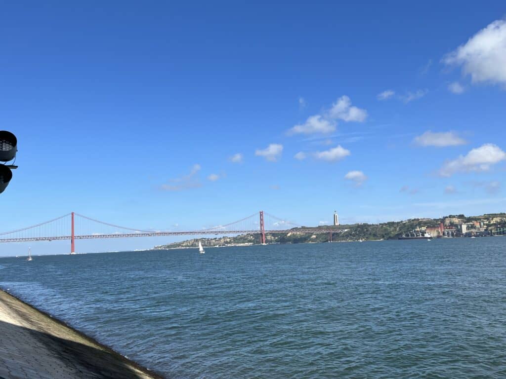 Red suspension bridge stretches across the Tagus River under a bright blue sky with a small sailboat on the water and the Cristo Rei statue visible in the distance. This scenic viewpoint is a highlight on a 3 day itinerary Lisbon and a gateway toward day trips from Lisbon.
