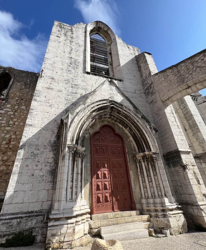 Carmo Convent in Lisbon shows a historic stone facade with a tall arched window and a red wooden door framed by detailed columns under a bright blue sky. Damaged during the 1755 earthquake, as evidenced by the visible fracture in the door frame, the partial ruins reflect the history explored on a 3 day itinerary Lisbon.
