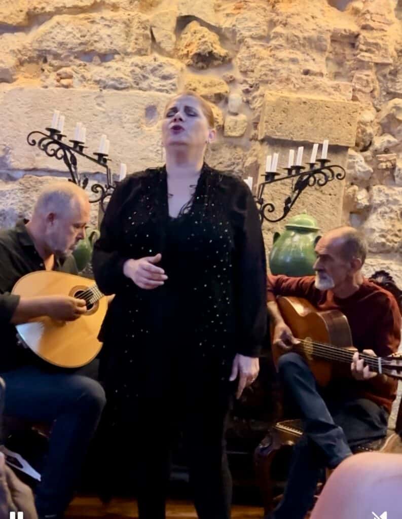 Woman dressed in black sings fado music while two musicians play Portuguese guitars beside her in an intimate Lisbon setting with a stone wall backdrop. Experiencing live fado is a cultural highlight to include in a 3 day itinerary Lisbon.