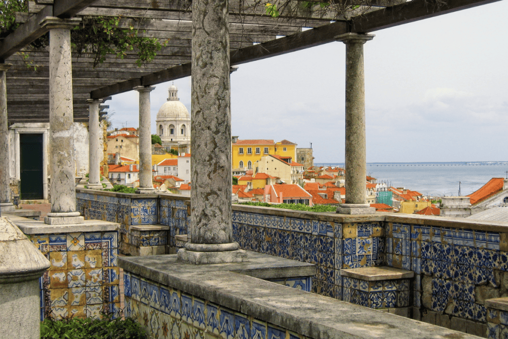 Scenic view from Miraduoro de Santa Luzia terrace with blue and white azulejo tiles and stone columns overlooks Lisbon’s rooftops and the Tagus River with a domed church in the distance. This viewpoint is a peaceful stop to include in a 3 day itinerary Lisbon.