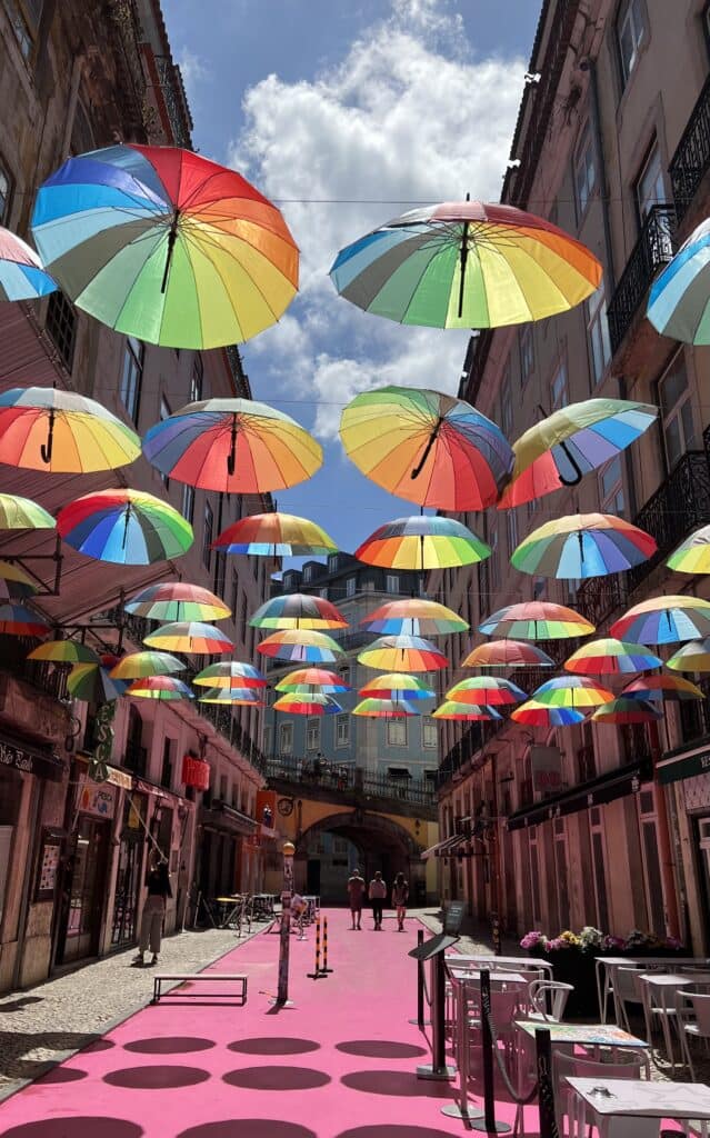 Street in Lisbon is decorated with colorful umbrellas hanging overhead casting shadows on a pink walkway below. Shops and cafes line the narrow street creating a lively atmosphere during a 3 day itinerary Lisbon.