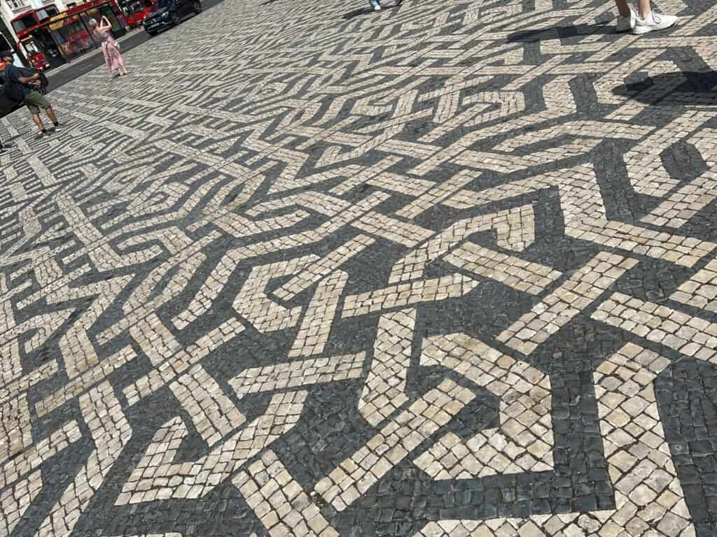 Black and white mosaic pavement in Lisbon forms an intricate geometric pattern across a large square with people walking in the distance. The traditional calçada portuguesa is a detail you will notice throughout a 3 day itinerary Lisbon.