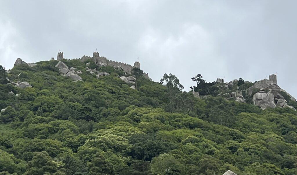 Moorish Castle walls stretch across a green hilltop in Sintra with stone towers rising above dense forest under a cloudy sky. This scenic viewpoint is a popular addition to a 3 day itinerary Lisbon when planning nearby day trips from Lisbon.