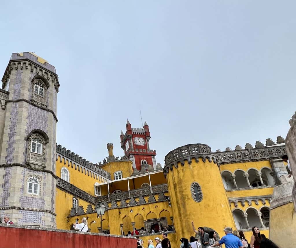Colorful Pena Palace in Sintra features bright yellow walls, a red clock tower, and detailed architectural elements with visitors exploring the courtyard. This fairytale castle is a must see stop when planning a 3 day itinerary Lisbon with nearby excursions.