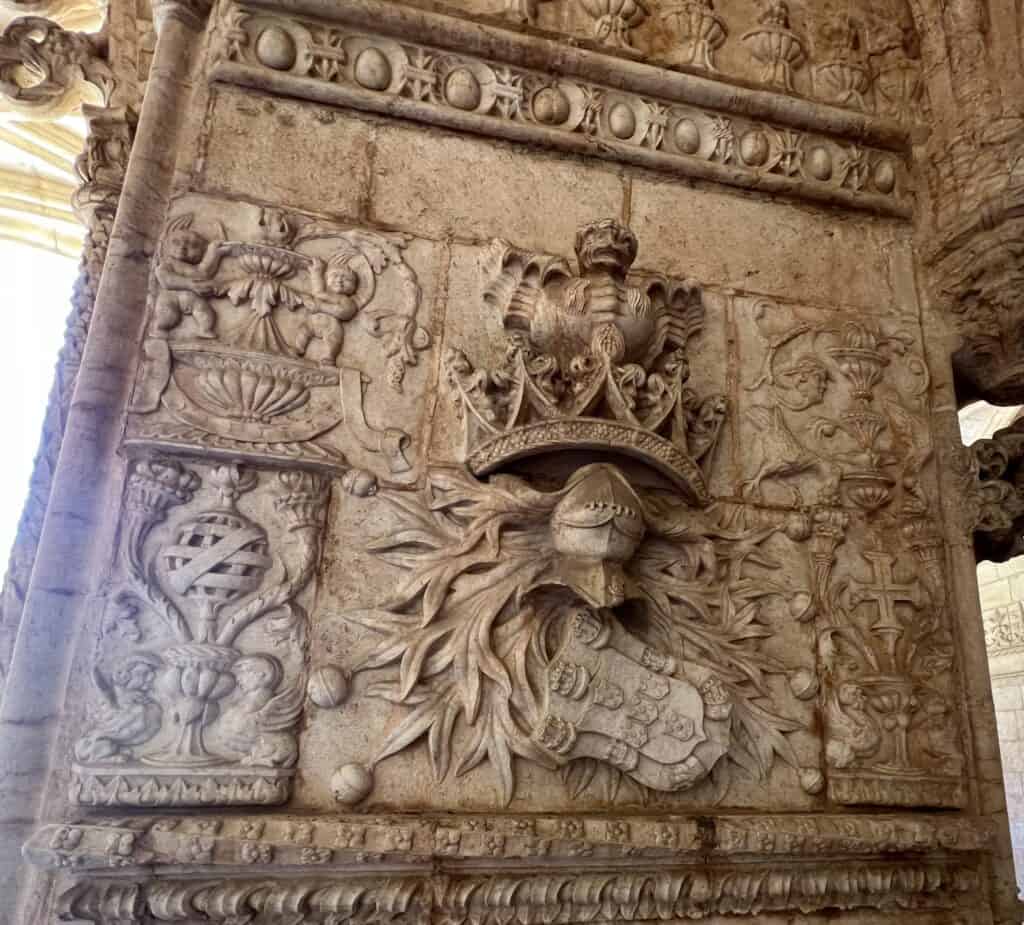 Close view of intricate stone carvings inside Jerónimos Monastery featuring a royal coat of arms surrounded by crowns vines and decorative symbols, including the armillary sphere. The detailed relief highlights the craftsmanship typical of Manueline architecture Lisbon.
