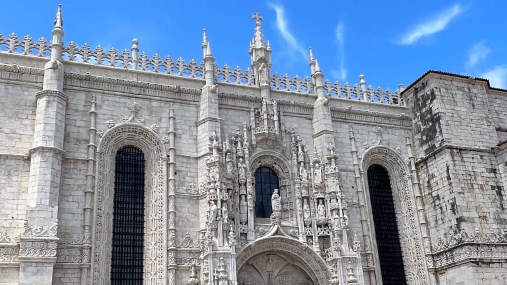 Ornate limestone facade of Jerónimos Monastery in Belém district Lisbon with tall arched windows and elaborate sculpted figures surrounding the entrance. The intricate carvings and decorative spires highlight the distinctive Manueline architecture Lisbon is known for.