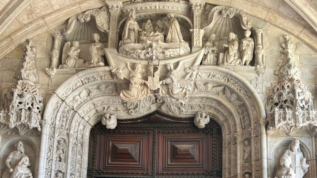 Elaborately sculpted entrance portal of Jerónimos Monastery filled with statues arches and religious figures above large wooden doors. The ornate facade is a defining example of Manueline architecture Lisbon.