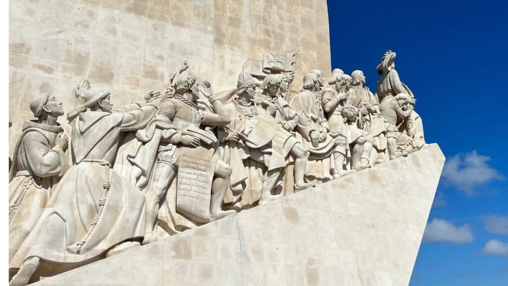 Sculpted explorers sailors and missionaries stand in a dramatic row along the Monument to Discoveries in Belém district Lisbon. The figures face the Tagus River celebrating leaders from the Age of Discovery Lisbon.