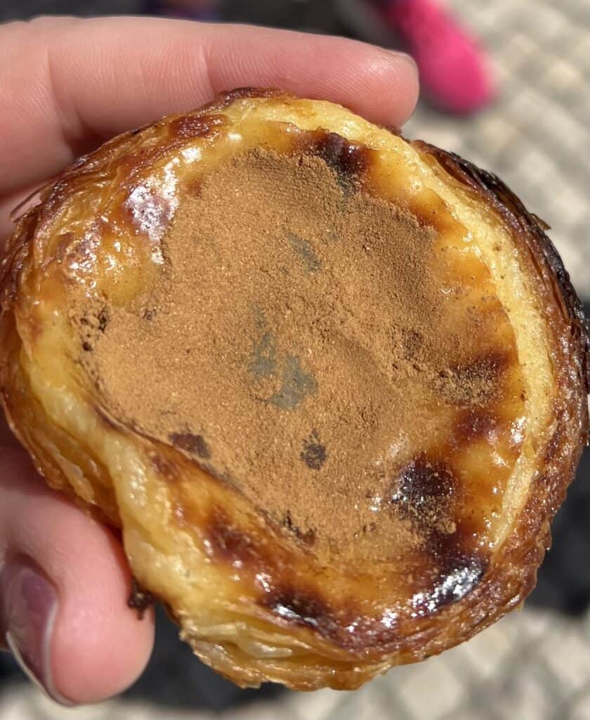 Pastel de nata Lisbon topped with a generous layer of cinnamon resting in a crisp golden pastry shell. The iconic custard tart is a must try dessert when visiting Pastéis de Belém in Belém district Lisbon.