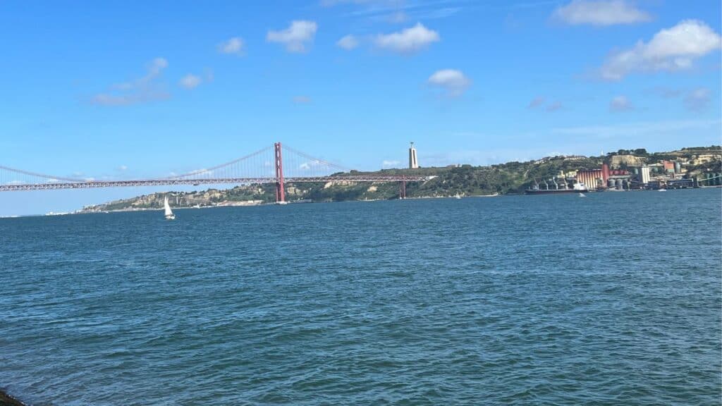 Wide view across the Tagus River toward the red suspension bridge known as the 25 de Abril Bridge and shoreline near Belém district Lisbon. Sailboats move across the water in the historic maritime setting linked to the Age of Discovery Lisbon. The statue in the distance is Cristo Rei