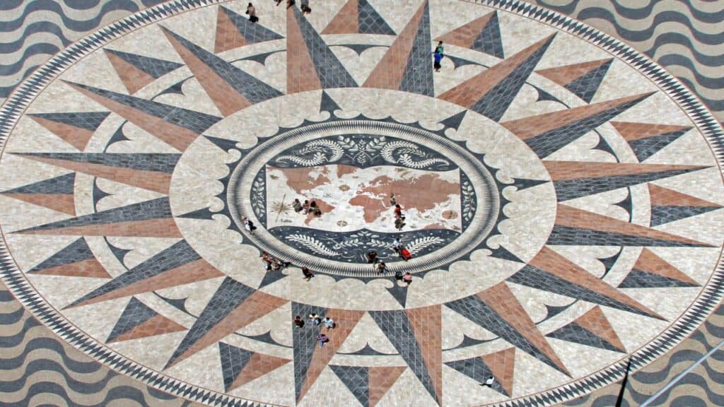 Aerial view of the large compass rose mosaic with a world map at its center near the Tagus River in Belém district Lisbon. The map shows Portuguese exploration routes. Visitors walk across the patterned stone design connected to the Age of Discovery Lisbon.