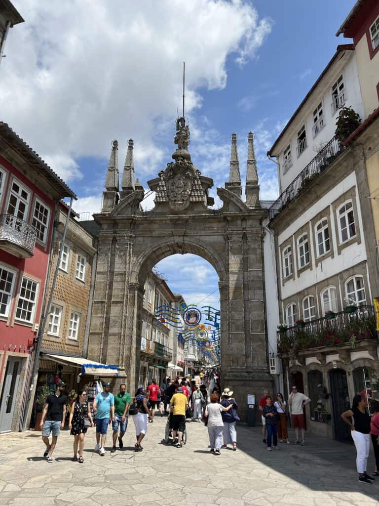 An ornate stone arch frames a lively pedestrian street lined with colorful buildings and festive decorations hanging overhead. Walking through historic streets like this is a classic experience when planning how to spend 3-5 days in Porto.