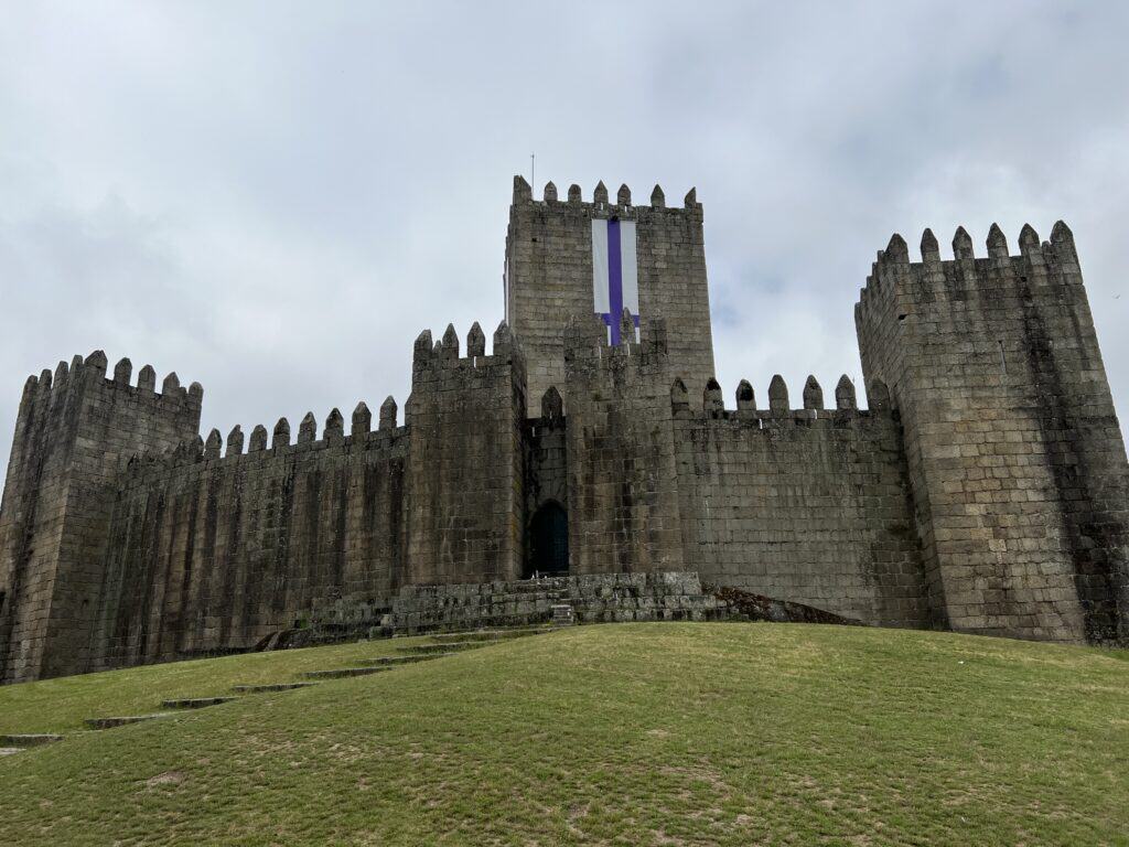Guimarães Castle, a medieval stone castle with tall towers and crenellated walls stands on a grassy hill under a cloudy sky. Visiting Guimarães is one of the day trips that can be taking during a 3-5 day Porto itinerary.