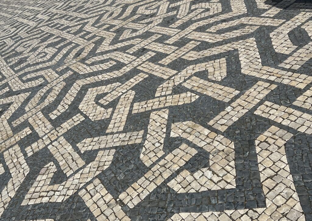 Black and white mosaic pavement in Lisbon forms an intricate geometric pattern across a large square with people walking in the distance. The traditional calçada portuguesa is a detail you will notice while in Portugal.