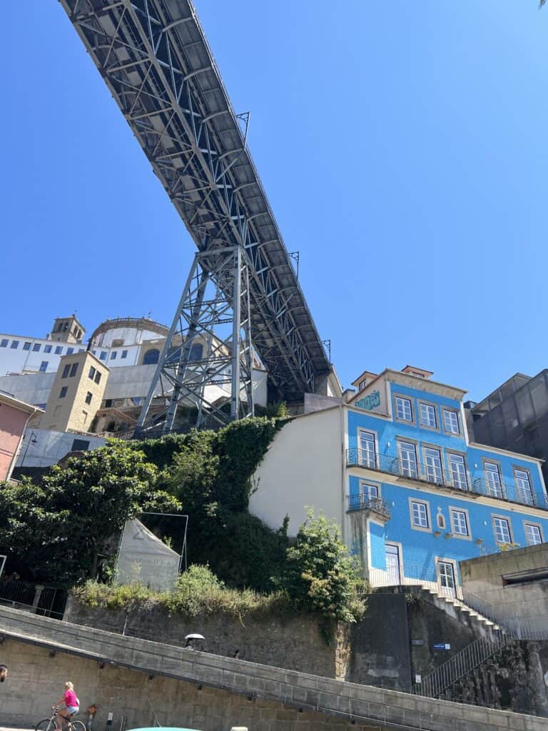 The underside of a bridge towers above a hillside with a bright blue tiled building and greenery along the Douro River. Views like this highlight how many days in Porto can be filled with scenic walks and riverfront sights.