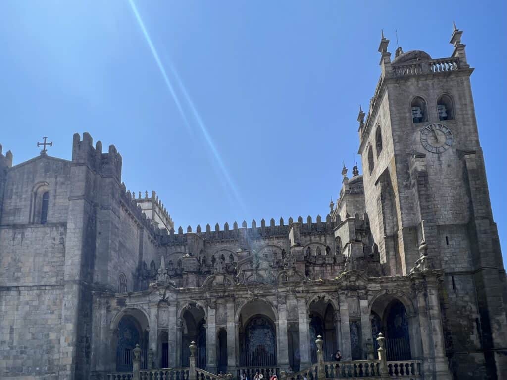 The ornate facade of Porto Cathedral rises with stone arches and a clock tower against a bright blue sky. Visiting this landmark is a must when planning how to spend 3-5 days in Porto.