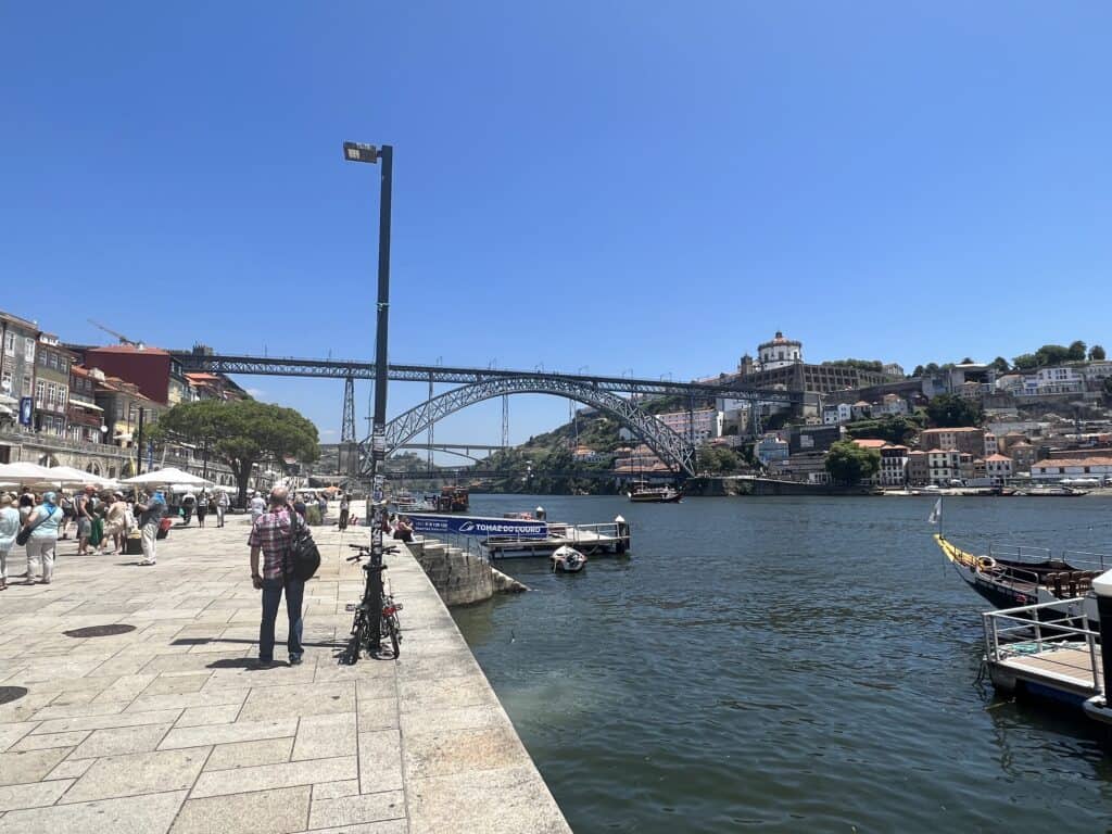 A sunny Ribiera promenade in Porto with people walking near the water and the Dom Luis I Bridge in the distance and Vila Nova de Gaia across the river. Strolling along the riverfront is one of the easiest things to do in Porto.