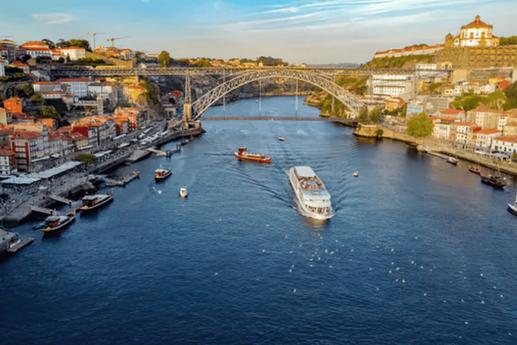 View over the Douro River in Porto with colorful riverside buildings and boats cruising beneath the Dom Luís I Bridge. This scenic perspective highlights a classic stop on a Porto itinerary with historic architecture lining both sides of the water.