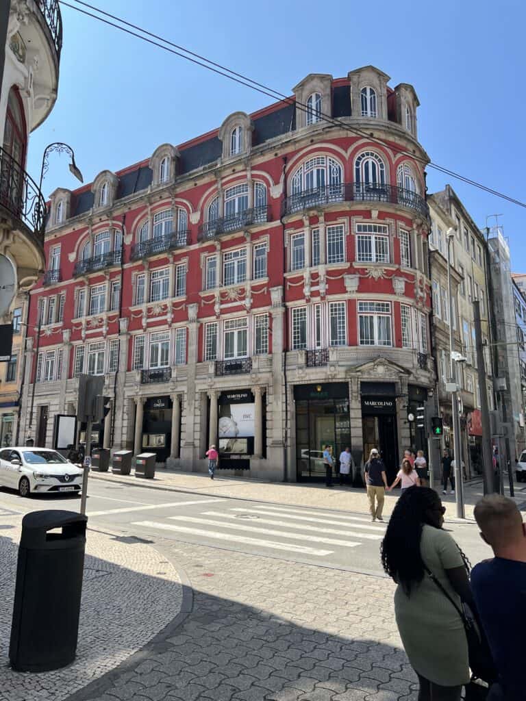 A curved red historic building with ornate Art Nouveau white trim and balconies sits on a sunny street corner in Porto as pedestrians cross the road. Strolling past architecture like this is one of the things to do in Porto when planning how to spend 3-5 days in Porto.