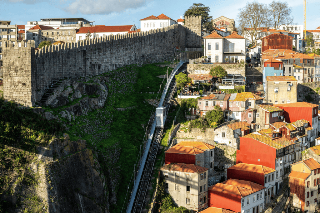 A steep hillside in Porto with colorful houses and a funicular railway climbing beside a medieval stone wall. Taking the funicular along this historic wall is a practical way to explore this hilly city.