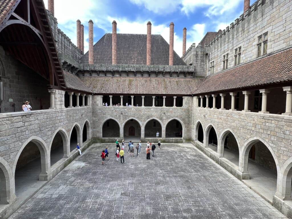 A stone courtyard at the Palace of the Dukes of Braganza is surrounded by arched galleries and red tiled roofs. Visitors gather in the open center while tall brick chimneys rise above the palace.